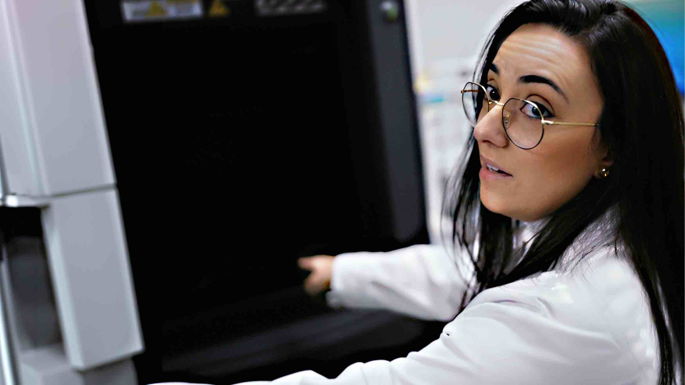 An individual in a lab coat and round glasses looking over their shoulder while working with laboratory equipment.